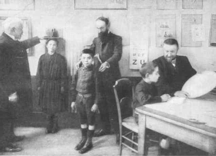 Binet examining a single boy with two adult observers in a classroom hung with educational posters.