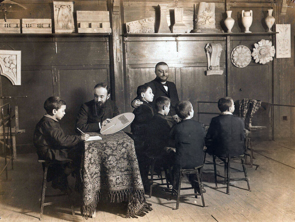 Alfred Binet at a table with five young boys in a classroom of plaster casts, conducting a group attention exercise.