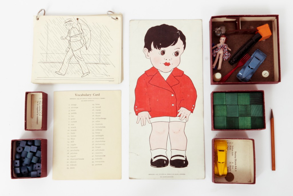 A vintage Stanford-Binet Intelligence Scale test kit, showing the classic illustrated boy figure, vocabulary card, picture card sets, blocks, and miniature objects used in the original assessment.